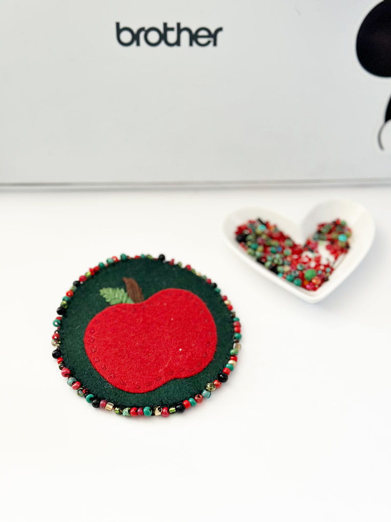 Felt apple brooch and beads in front of Brother ScanNCut cutting machine
