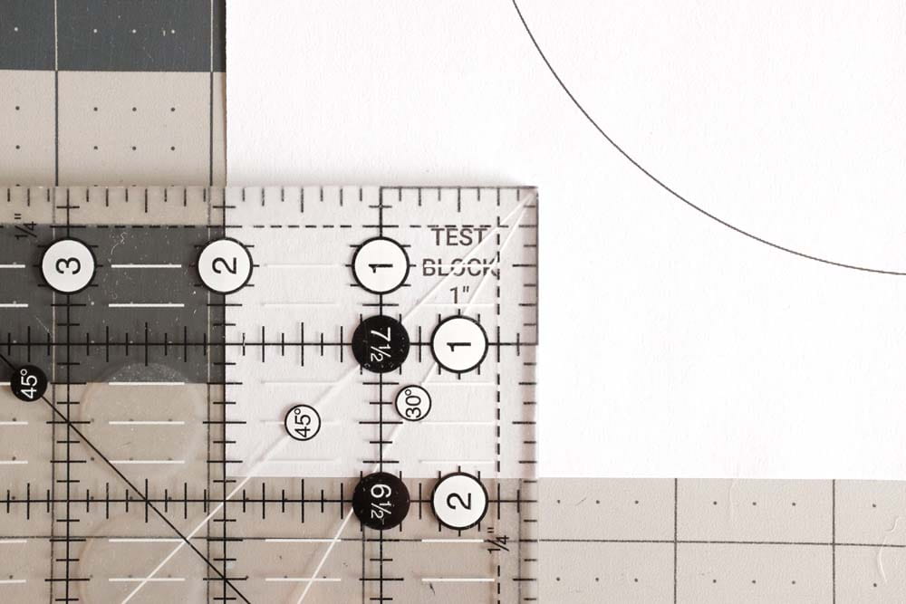 A close-up of a transparent quilting ruler placed on a cutting mat with measurement markings. The ruler displays various lines and angles, including 45 and 30-degree angles, along with labeled measurements for precision cutting.