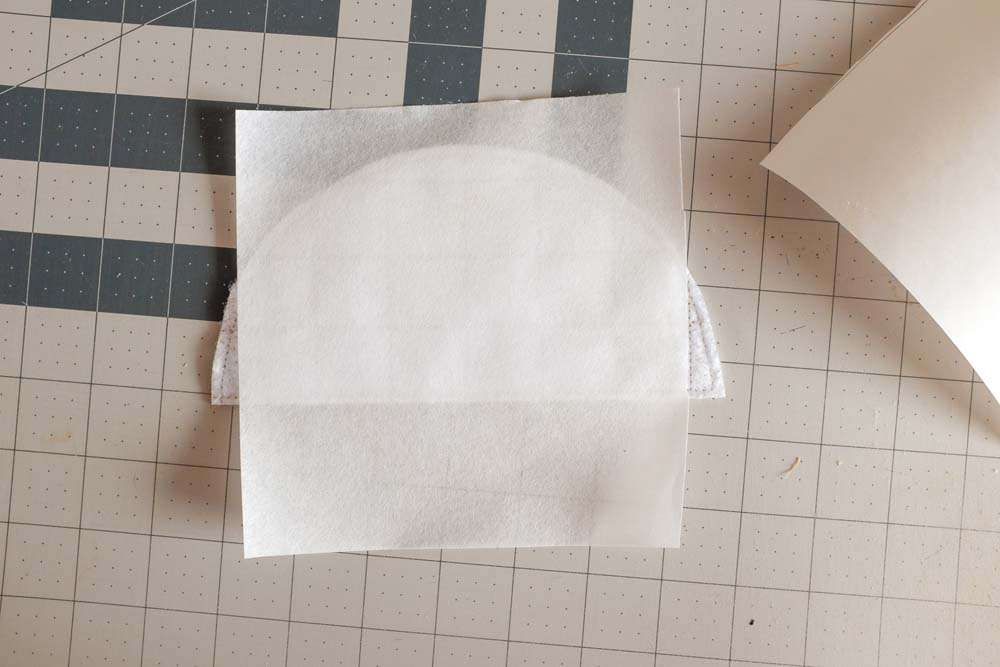 A semicircular fabric piece layered with batting and protective paper sheets, placed on a cutting mat, ready for assembly or further quilting steps.