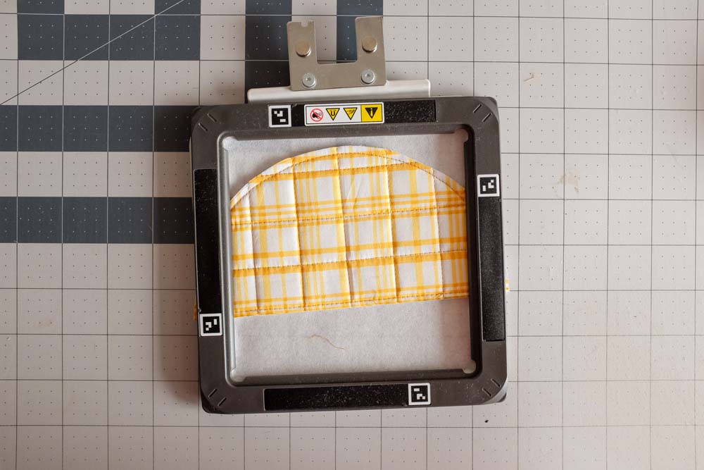 A semicircular quilted fabric in yellow plaid is positioned inside a black embroidery machine frame, layered with stabilizer for embroidery.