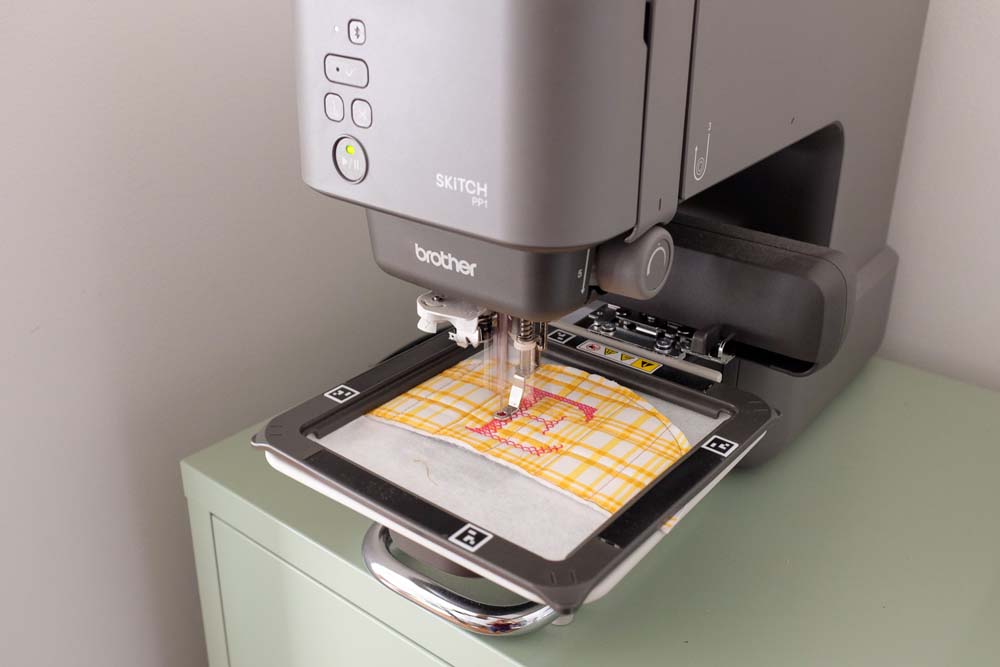 A Brother embroidery machine actively stitching a design onto the yellow plaid quilted fabric secured in the embroidery frame. The machine's screen and controls are partially visible.