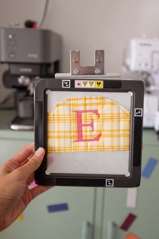 A completed embroidered yellow plaid semicircle is shown in the embroidery frame, featuring a bold letter "E" in pink thread at the center.