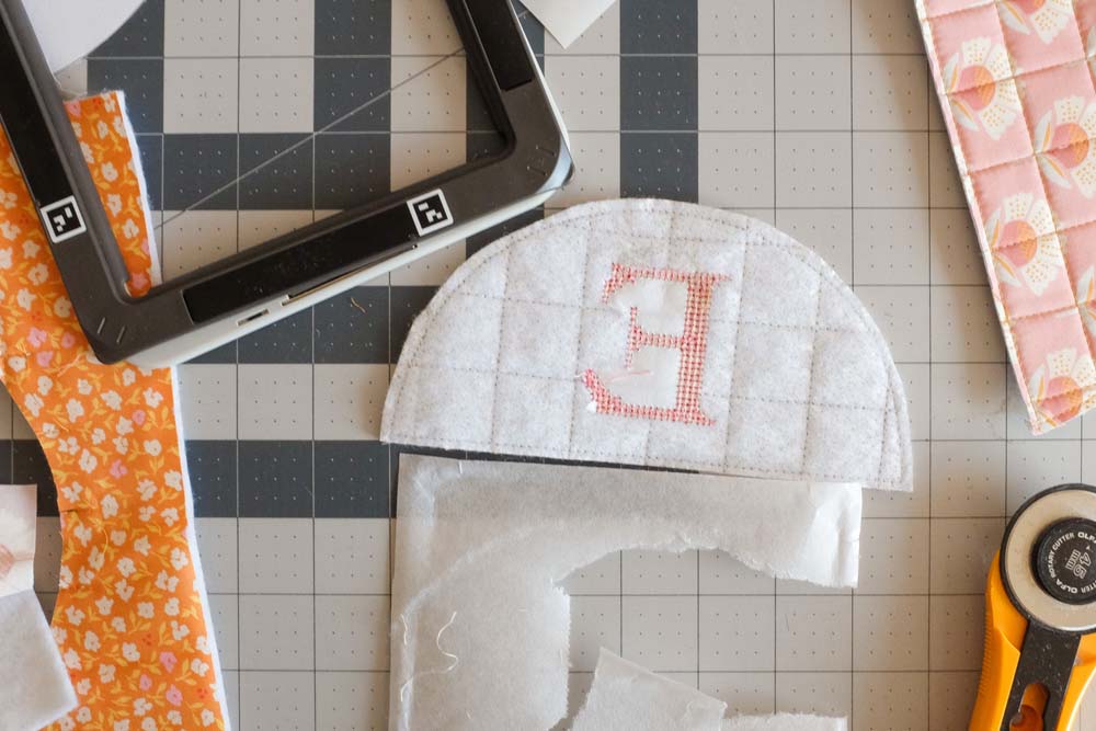 The reverse side of the embroidered yellow plaid semicircle, revealing the stabilizer backing and raw stitching on a cutting mat. A rotary cutter and fabric scraps are nearby.