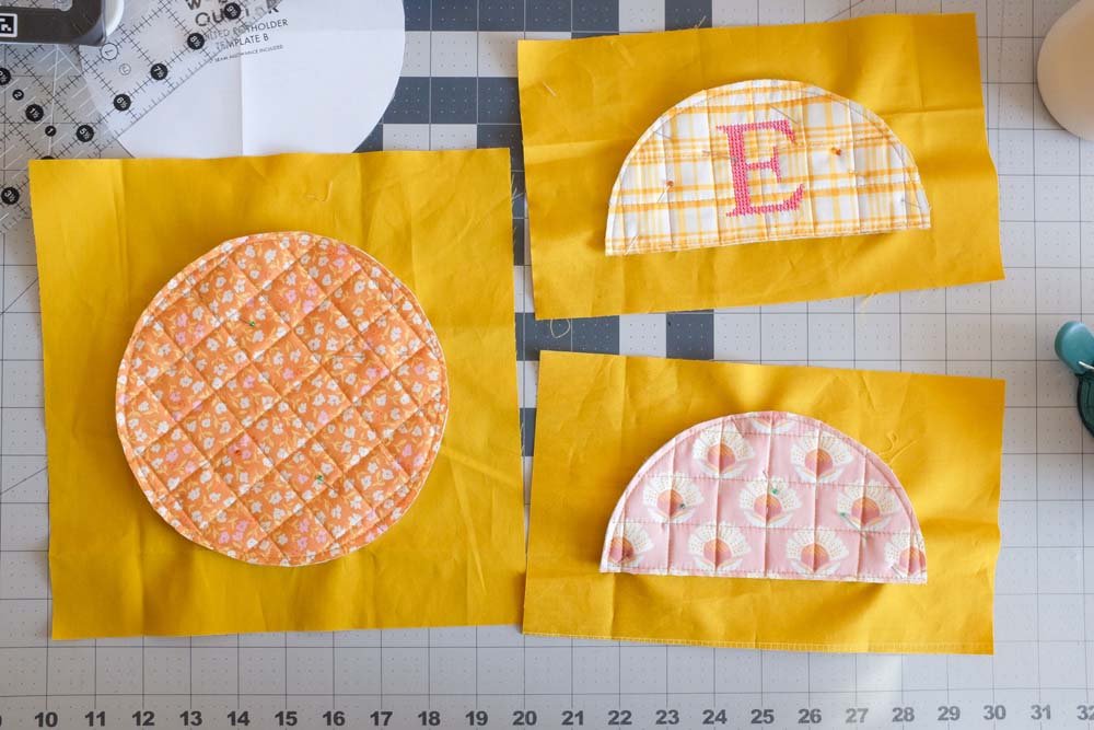 Three quilted fabric pieces—a circular orange floral piece, a semicircular pink floral piece, and a yellow plaid piece with an embroidered "E"—are placed on rectangular yellow fabric backgrounds.