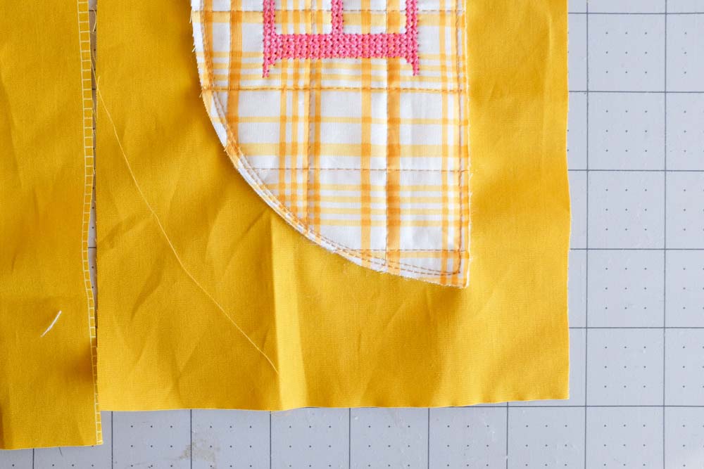 A close-up of the yellow plaid quilted semicircle with the embroidered "E" being stitched onto a yellow fabric background. The edge stitching is visible.