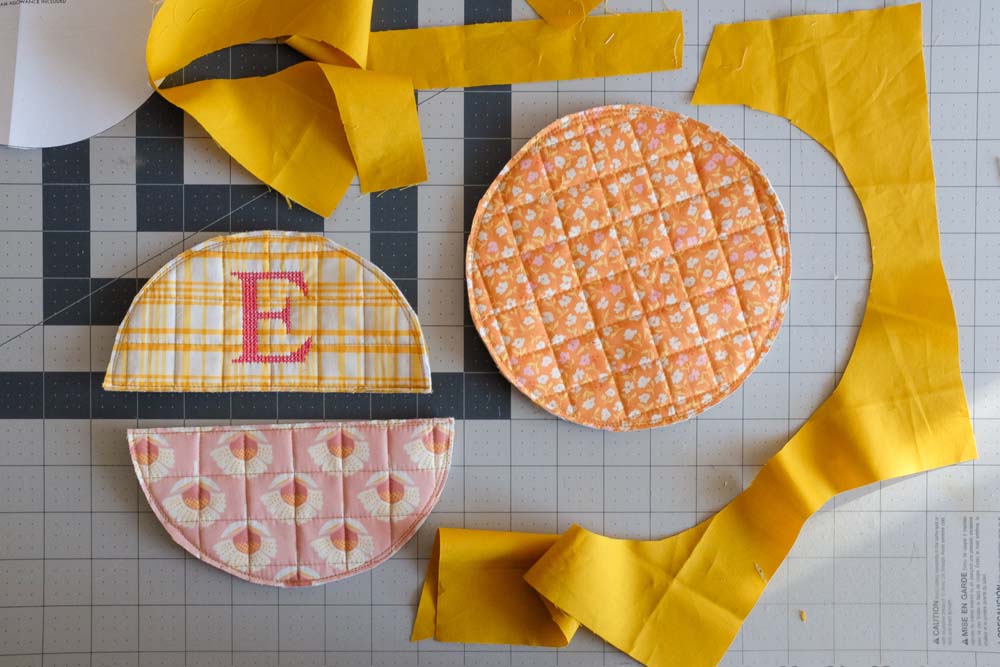 Three assembled quilted potholder pieces—a circular orange floral piece, a semicircular pink floral piece, and a yellow plaid piece with the embroidered "E"—are arranged on a cutting mat alongside yellow fabric strips.