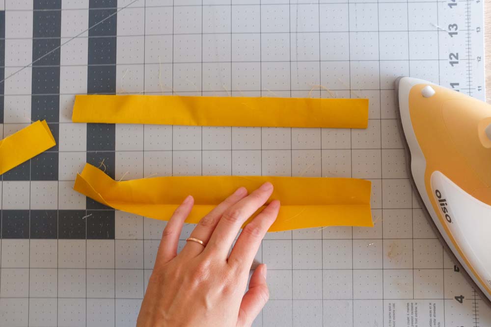 Yellow fabric strips being prepared for binding, with one strip folded lengthwise and partially pressed. A hand adjusts one strip, and an iron is visible on the cutting mat.