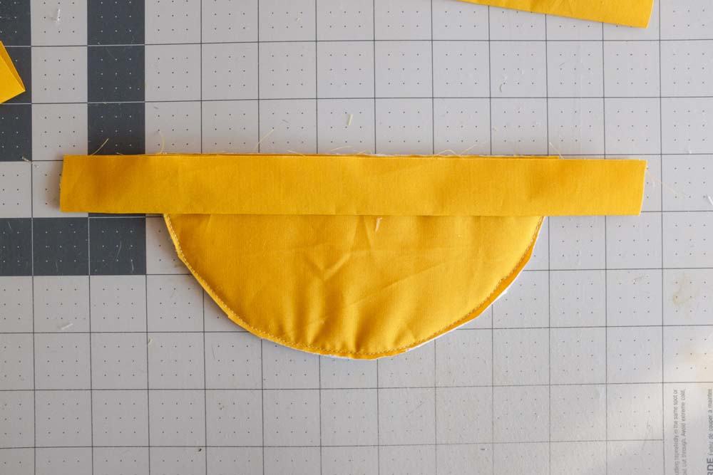 A yellow fabric semicircle is laid flat on a cutting mat with a rectangular yellow fabric strip positioned at the top edge, ready for sewing.
