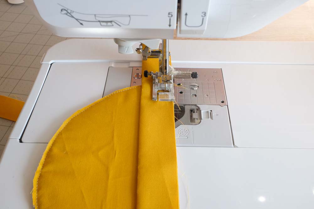 A sewing machine is stitching the yellow fabric strip to the yellow semicircle, showing the progress of attaching the binding.