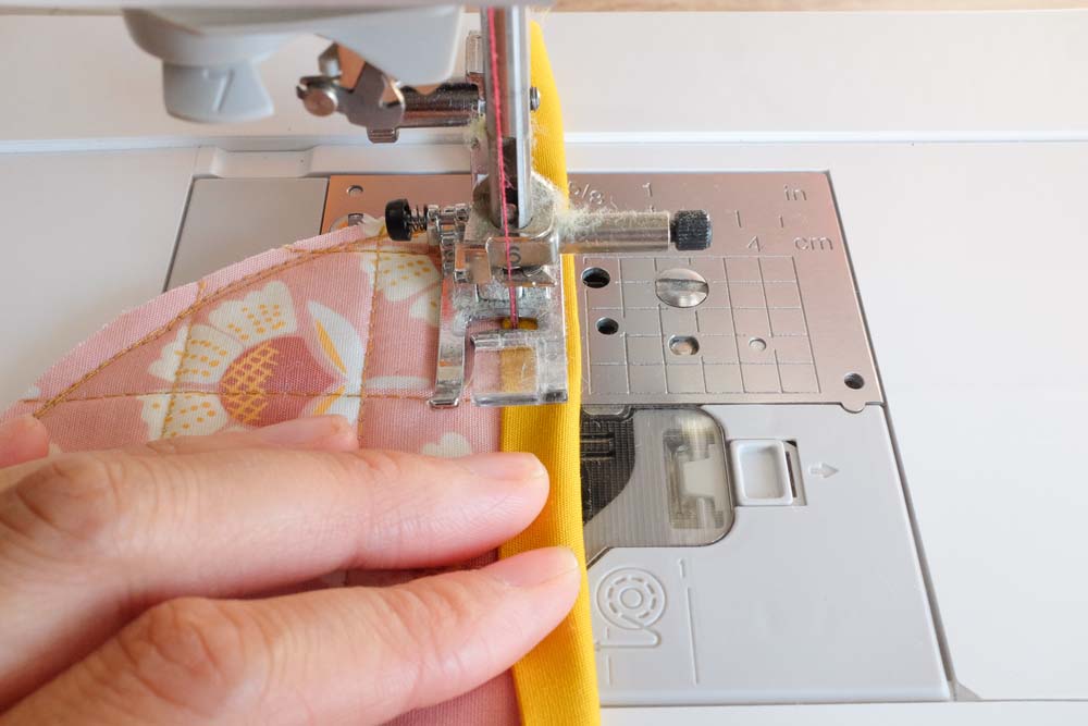 A close-up of a sewing machine needle stitching the yellow binding onto the pink floral semicircle.