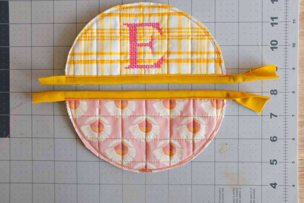 Two semicircular quilted fabric pieces, one yellow with a monogrammed "E" and one pink floral, each with yellow fabric bindings sewn to the straight edges.