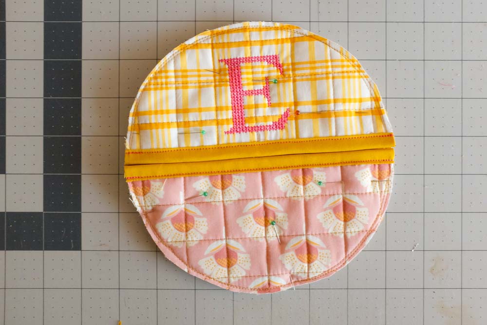 The final circular potholder design with the yellow monogrammed "E" semicircle on top and the pink floral semicircle at the bottom, both with yellow bindings and securely pinned in place.