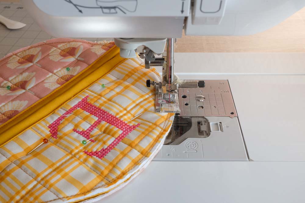 A sewing machine stitching the combined quilted layers together, finalizing the assembly of the circular potholder design.