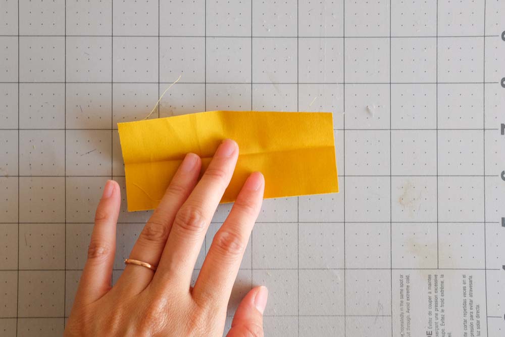 A rectangular strip of yellow fabric being prepared, pressed flat with visible fold lines, and held by a hand against a grid cutting mat.