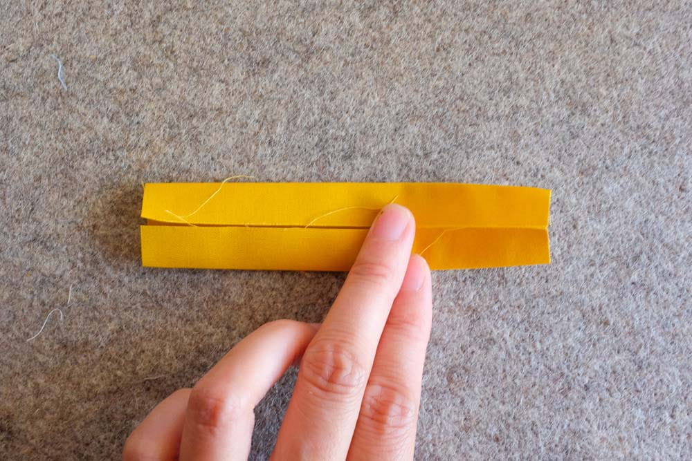 A strip of yellow fabric being folded and pressed with a finger on a wool ironing mat, showing neat fold lines for a binding loop.