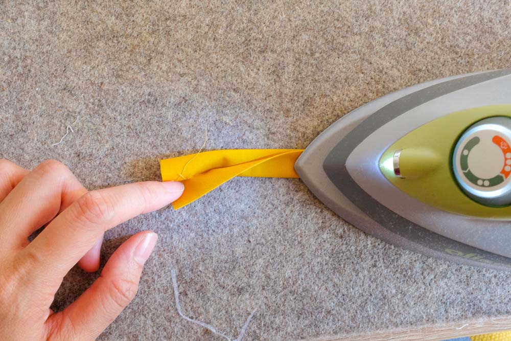 A close-up of an iron pressing a folded yellow fabric strip on a wool ironing mat, with fingers holding the fabric in place for precision.