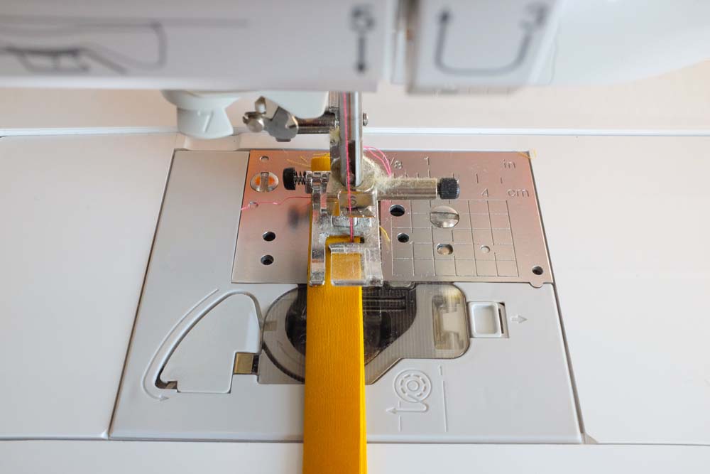 A close-up of a sewing machine needle stitching along the center of a folded yellow fabric strip, securing the layers together.