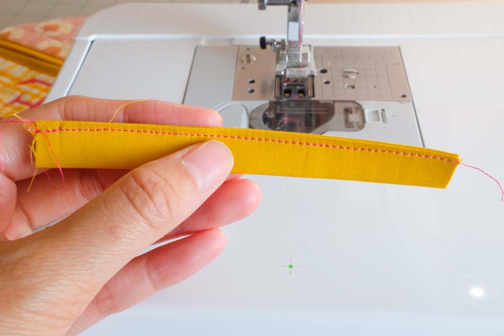 A hand holding a stitched yellow fabric strip, showing neat pink stitching along the edge.