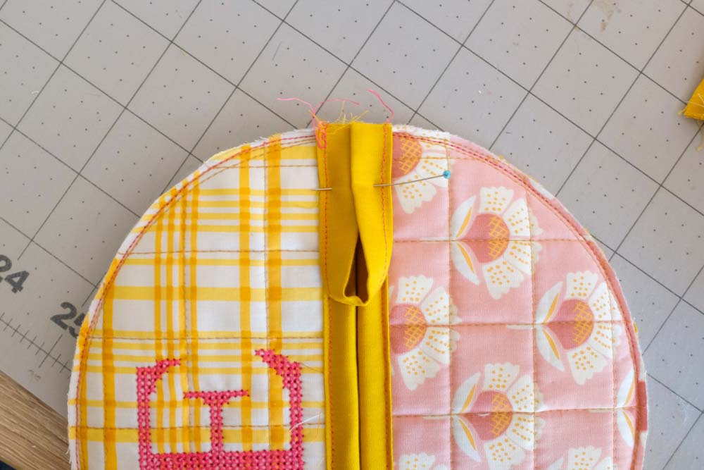 A quilted circular fabric piece with yellow plaid and pink floral patterns. A folded yellow strip is pinned at the top as a loop for hanging.