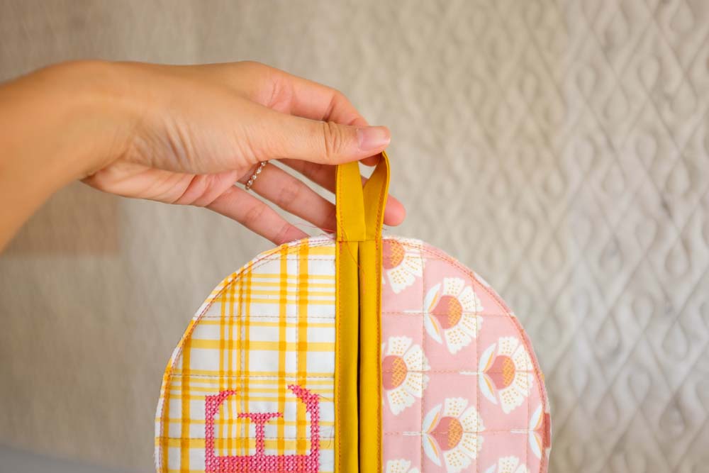 A completed potholder with quilted floral and plaid fabric sections, yellow edging, and a hanging loop, held up by a hand in front of a quilted background.