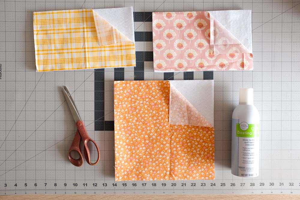Three layers of fabric are laid out on a cutting mat, including printed fabric in yellow plaid, pink floral, and orange floral patterns, each layered with white batting. A pair of scissors and a can of quilt basting spray are placed nearby.