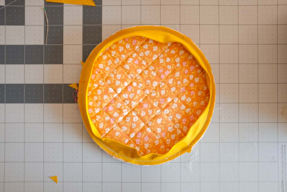 A circular quilted fabric piece with bright orange and yellow floral patterns, pinned with yellow binding fabric around its edges.