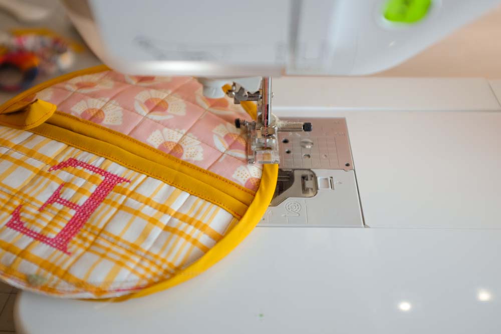 A sewing machine stitching yellow binding onto a quilted circular piece with a pink and yellow floral design and cross-stitch embroidery.