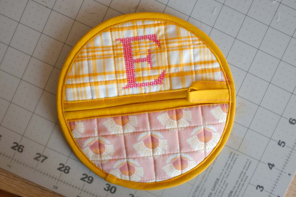The finished quilted circular potholder, featuring yellow plaid and floral fabrics with a pink cross-stitched letter "E" and bright yellow binding.
