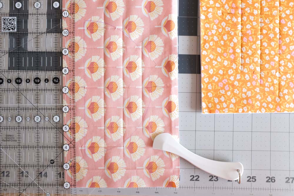 A close-up of a pink floral fabric with grid quilting lines drawn and stitched. A transparent quilting ruler and a white fabric marking tool are placed beside it on the cutting mat.
