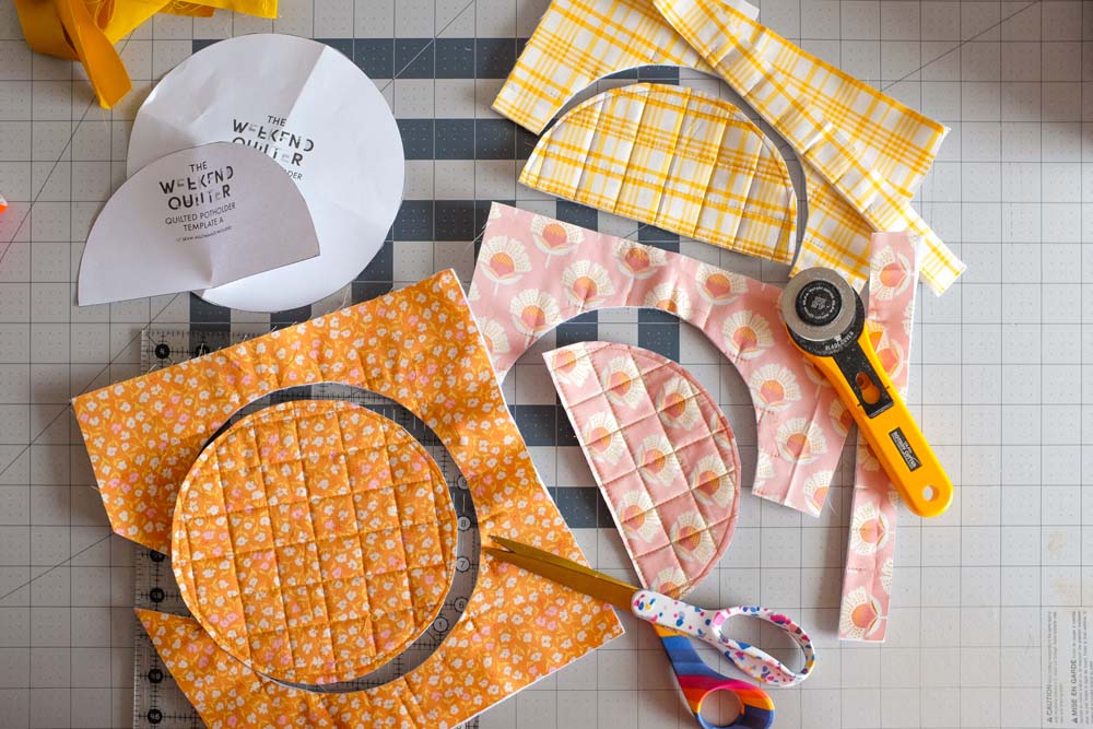 Various fabric pieces, including semicircles and strips, cut from quilted fabric panels in yellow plaid, pink floral, and orange floral designs. A rotary cutter, scissors, and templates are arranged alongside the pieces on a cutting mat.