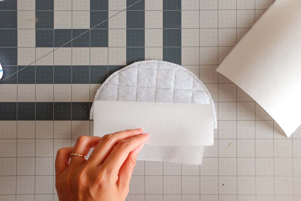 A hand holding a piece of batting layered with quilted fabric, partially wrapped with parchment paper or protective fabric sheets to prepare for pressing or layering.