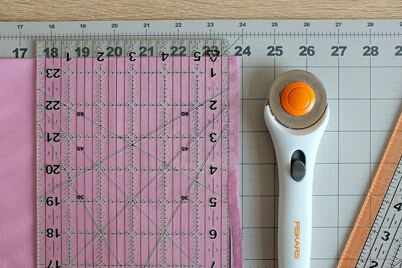 Pink fabric on cutting board with ruler on top and rotary cutter next to it