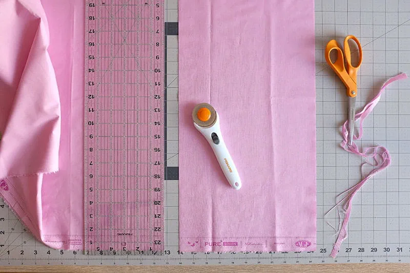 Ruler on pink fabric on a cutting board with rotary cutter and scissors.