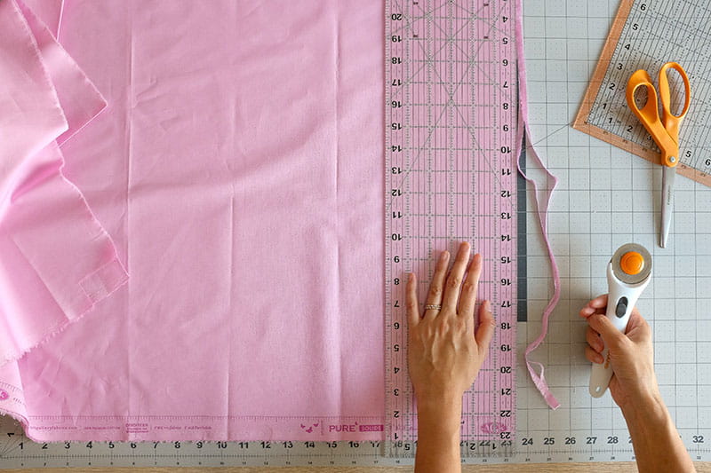 Person holding ruler against pink fabric and using a rotary cutter to trim edges.