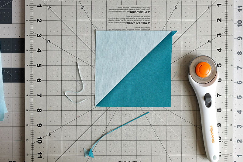 Blocked light blue and teal fabric to make a square with triangles next to a rotary cutter on a cutting board