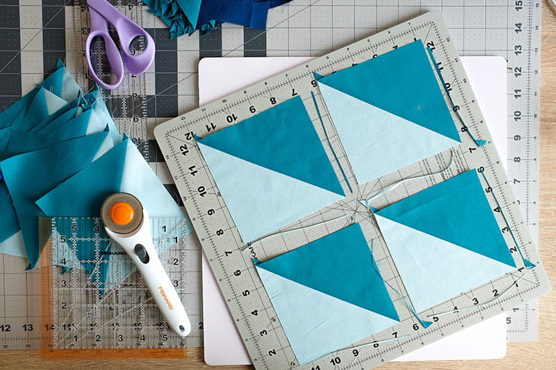 Four half square triangles on a cutting board next to completed fabric squares with scissors and rotary cutter on left.