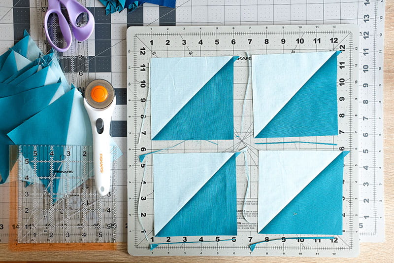 Four half square triangles on a cutting board with trimmings next to completed fabric squares with scissors and rotary cutter on left. 