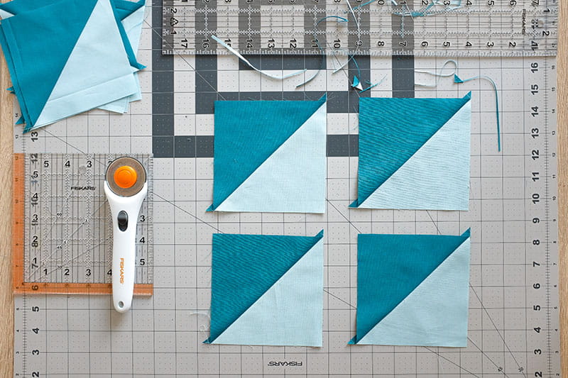 Four half square triangles on a cutting board with rulers above and to the left and a rotary cutter on left