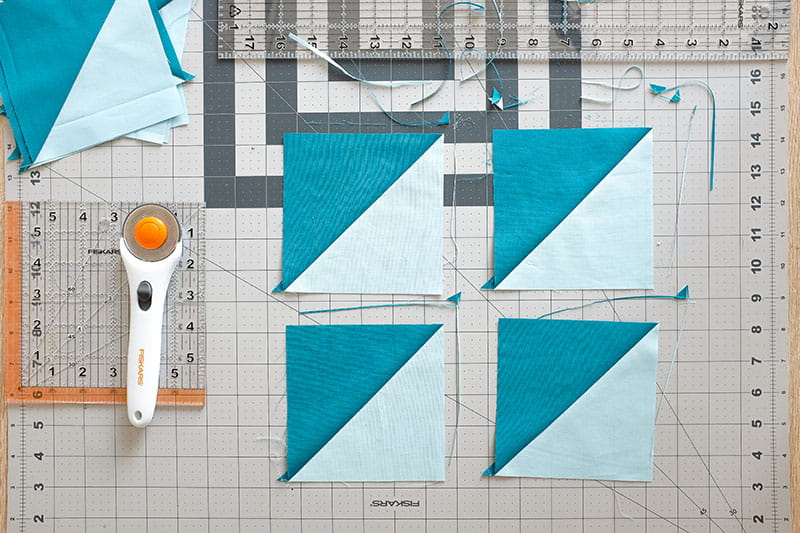 Trimmed half square triangles in light blue and teal on a cutting board with quilting ruler and rotary cutter