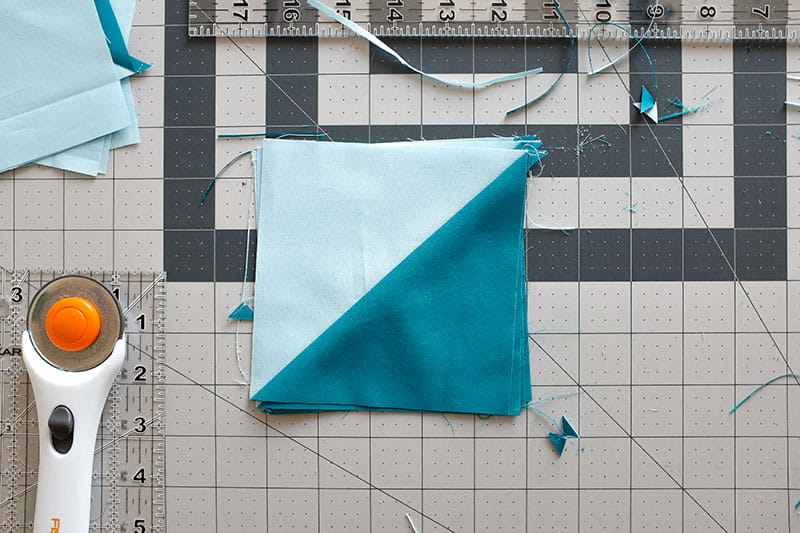 Light blue and teal half square triangle on a cutting board with rotary cutter and quilting ruler.