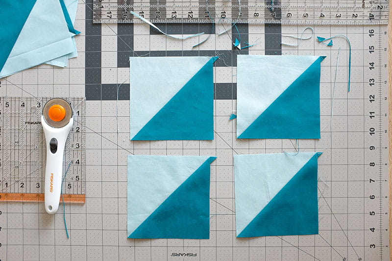 Four half square triangles on a cutting board with quilting rulers and rotary cutter.