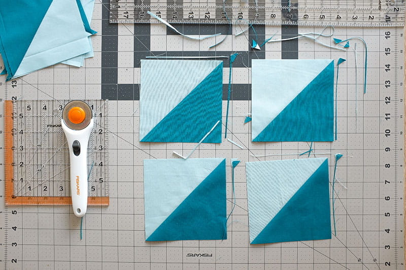 Four half square triangles with visible trimmings on a cutting board with rotary cutter and quilting ruler