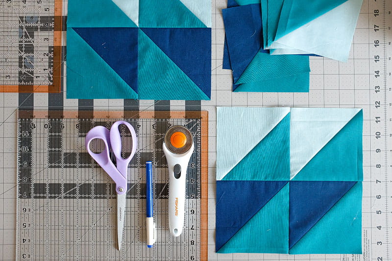 Blocked half square triangles with light blue and teal and royal blue and teal on a cutting board next to scissor, rotary cutter and fabric pen.