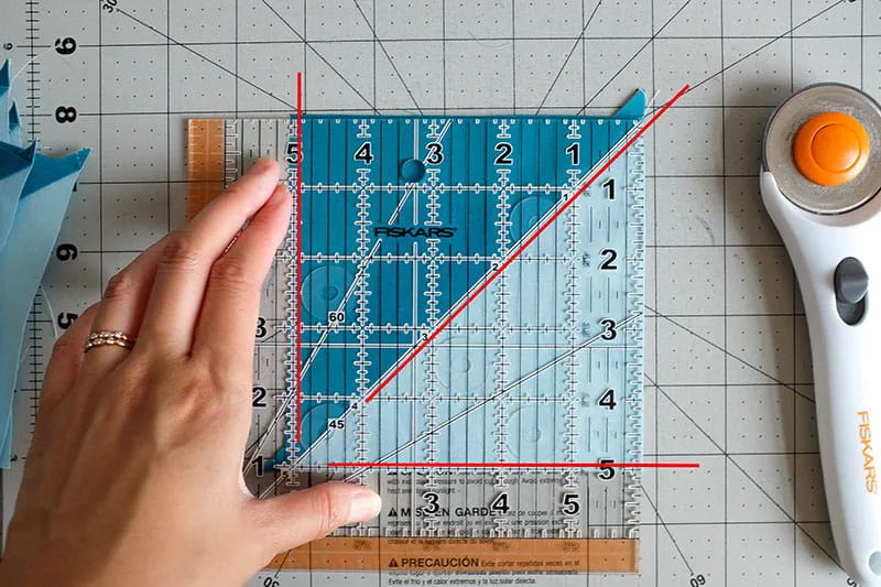Person lining up quilting ruler to align two fabrics of half square triangle on a cutting board.