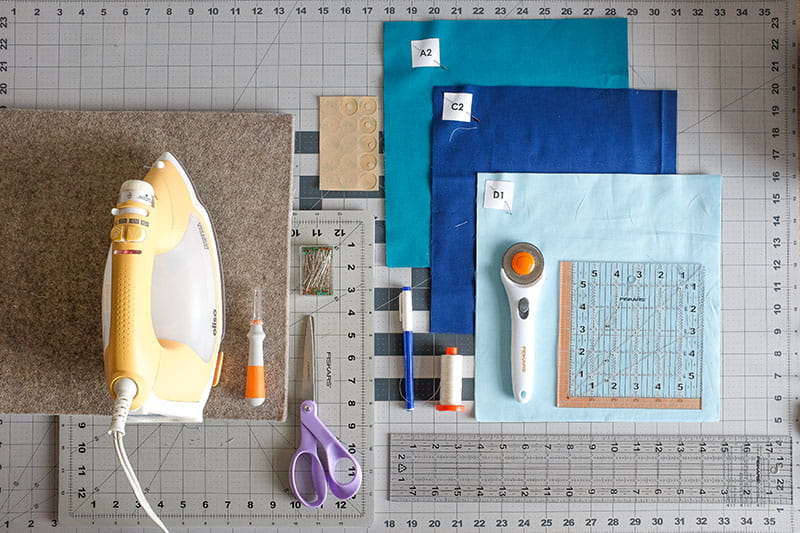 Supplies for creating half square triangles including iron, fabrics, rotary cutter, scissor, fabric pen and quilting rulers