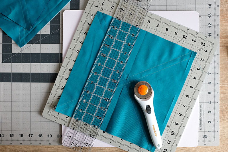 Large square of teal fabric on a cutting board with quilting ruler on top and fabric cutter.