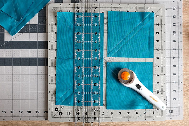 Large square of teal fabric cut into one rectangle and two squares on a cutting board with quilting ruler on top and fabric cutter.