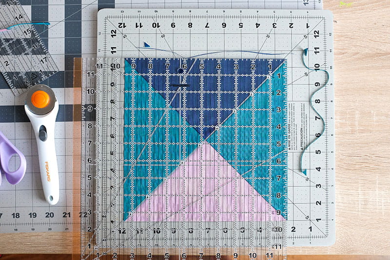 Fully aligned quarter square triangle piece with a quilting ruler on top next to a rotary cutter.