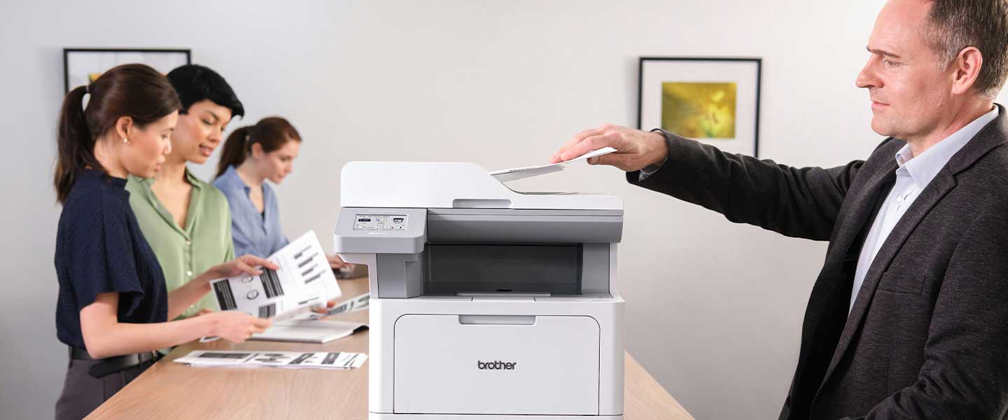 Man printing document on Brother MFCL5715DW printer in an office with coworkers on opposite side of table.