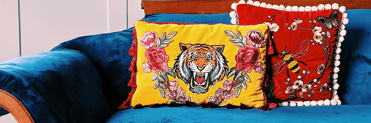 Tiger embroidery design on a yellow pillow and insect and flowers embroidery on a red pillow, both on a blue velvet couch. 